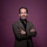 Portrait von Dr. Benny Pock im brauen Anzug vor weinrotem Hintergrund.