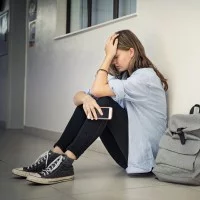 Trauriges Mädchen mit einem Smartphone in der Hand, das auf dem Boden im Flur einer Schule sitzt und den Kopf hält.