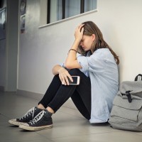 Trauriges Mädchen mit einem Smartphone in der Hand, das auf dem Boden im Flur einer Schule sitzt und den Kopf hält.