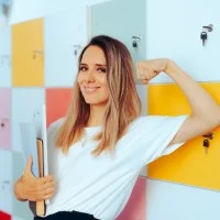 Glückliche Lehrerin vor Schließfächern, die ihren starken Bizeps zeigt und dabei Dokumente in der Hand trägt.