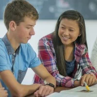 Vier Schülerinnen und Schüler sitzen am Tisch und lernen zusammen. Im Fokus ist eine Schülerin asiatischer Abstammung, die mit dem Stift auf ein Buch zeigt und lächelt.