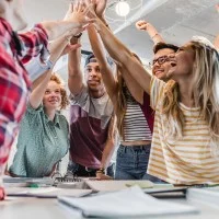Sechs Jugendliche unterschiedlicher Herkunft und Geschlecht stehen an einem Tisch auf dem Hefte liegen und machen einen Huddle.