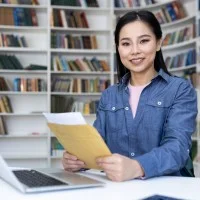 Junge Frau mit Zeugnis sitzt vor einem Buchregal am Laptop