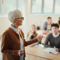 Eine alte Lehrerin steht im Klassenzimmer vor einer Gruppe von Schülerinnen und Schülern und erklärt den Unterricht. Die Lernenden hören aufmerksam zu und beteiligen sich am Gespräch.