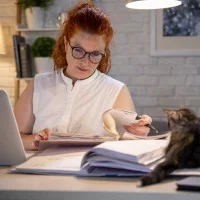 Eine Lehrerin arbeitet zuhause am Laptop und macht sich Notizen in ein Heft. Auf dem Schreibtisch liegt eine Katze.
