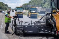 Ein Asphaltbauer, gefolgt von zwei Straßenwalzen, verbaut Flüsterasphalt bei der Sanierung der Autobahn A3 in Nordrhein-Westfalen.