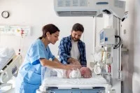 Eine Gesundheits- und Kinderkrankenpflegerin kontrolliert den Gesundheitszustand eines Neugeborenen. Daneben steht der Vater des Babys, der mit seiner Hand das Baby streichelt.