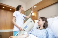 Eine Gesundheits- und Krankenpflegerin überprüft die Einstellung des Tropfes einer Patientin. Diese liegt im Krankenzimmer in ihrem Krankenbett.