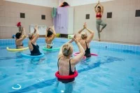 Eine Masseurin und medizinische Bademeisterin leitet vom Beckenrand aus eine Gruppe von fünf Frauen im Wasser zu gemeinsamen Yoga-Übungen an. Die Frauen sind im Schwimmbecken durchführen und haben eine Poolnudel und dem Bauch gelegt. 