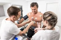 Eine medizinische Technologin und ein Technologe für Funktionsdiagnostik befestigen Messelektroden am nackten Oberkörper eines Patienten. 