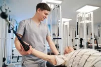 Ein Physiotherapeut unterstützt einen älteren Herrn in einem Fitnessraum dabei, seine Kraft in den Armen zu trainieren. 