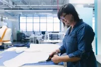 Eine bekleidungstechnische Assistentin schneidet auf einem Tisch in einer hellen Halle eine Entwurfsskizze zu einen Musterschnitt zurecht. 