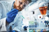 Ein chemisch-technischer Assistent untersucht in Schutzkleidung im Labor eine Probe mit einer hellblauen Reagenz. Weitere Reagenzien stehen auf dem Tisch.