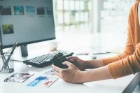 Hände halten eine Kamera vor einem Computerbildschirm. Auf dem Tisch befinden sich Fotos, eine Tastatur und ein Stiftehalter.