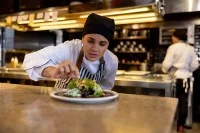 Eine gastronomische Assistentin garniert eine Speise in der Großküche. Im Hintergrund arbeitet eine weitere Person aus dem Betrieb.