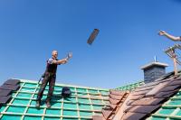 Ein Dachdecker wirft seinem Kollegen auf dem Dach einen Ziegel zu.