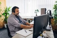 Ein übergewichtiger Mann mit einem Headset auf dem Kopf sitzt im Büro an seinem Schreibtisch und tippt etwas in die Computertastatur. 