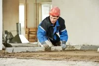 Ein Estrichleger verteilt Estrichmörtel mit einer Abziehlatte auf dem Boden eines Gebäudes.