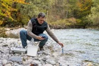 Ein umweltschutztechnischer Assistent hockt neben einem Flusslauf und hält ein Messgerät in das Wasser.