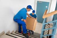 Zwei Fachkräfte für Möbel-, Küchen- und Umzugsservice transportieren einen Umzugskarton mit einer Sackkarre die Treppe hinunter.
