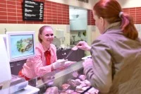 Eine Fachverkäuferin im Lebensmittelhandwerk bietet einer Kundin an der Fleischtheke ein Stück Wurst zum Probieren an.