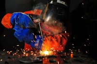 Ein Schweißer in orangener Schutzkleidung, blauen Handschuhen und Helm, der etwas schweißt. Der Hintergrund ist schwarz und Funken sprühen.