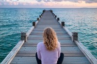 Eine Frau sitzt mit dem Rücken zum Betrachter auf einem Bootssteg und blickt zum Wasser sowie Horizont.