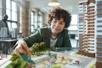 Eine Frau mittleren Alters sitzt in einem Büro vor einem Modell und berührt dieses mit der rechten Hand. Auf dem Modell ist ein Wohnblock, eine Freizeitanlage, eine Grünfläche und eine Straße gebaut.