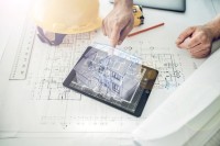 Auf einem Tisch liegt eine Bauzeichnung, auf der wiederum ein Tablet mit einem Hologramm eines Hauses sowie ein Lineal, Helm und Stift liegen. Eine Hand berührt das Hologramm.