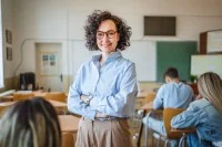 Eine Lehrerin steht mit verschränkten Armen in ihrer Klasse. Sie wirkt ruhig, gelassen und kompetent.