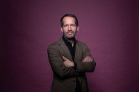 Portrait von Dr. Benny Pock im brauen Anzug vor weinrotem Hintergrund.