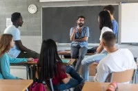 Ein Berufsschullehrer lehnt am Pult und diskutiert mit einigen Schülerinnen und Schülern.