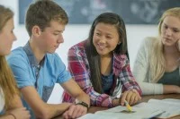 Vier Schülerinnen und Schüler sitzen am Tisch und lernen zusammen. Im Fokus ist eine Schülerin asiatischer Abstammung, die mit dem Stift auf ein Buch zeigt und lächelt.