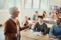 Eine alte Lehrerin steht im Klassenzimmer vor einer Gruppe von Schülerinnen und Schülern und erklärt den Unterricht. Die Lernenden hören aufmerksam zu und beteiligen sich am Gespräch.