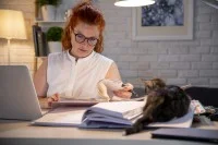 Eine Lehrerin arbeitet zuhause am Laptop und macht sich Notizen in ein Heft. Auf dem Schreibtisch liegt eine Katze.