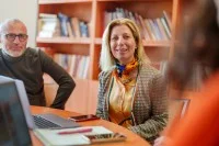 Drei Personen sitzen in einem Büro. Auf dem Tisch sind Schreibutensilien, ein Laptop und ein Handy.