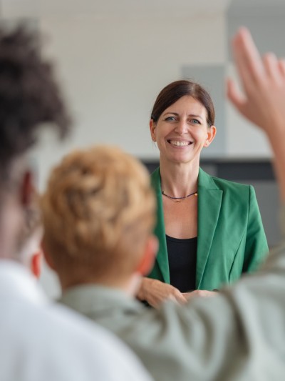 Eine lächelnde Lehrerin steht vor ihrer Klasse, ein Schüler meldet sich.