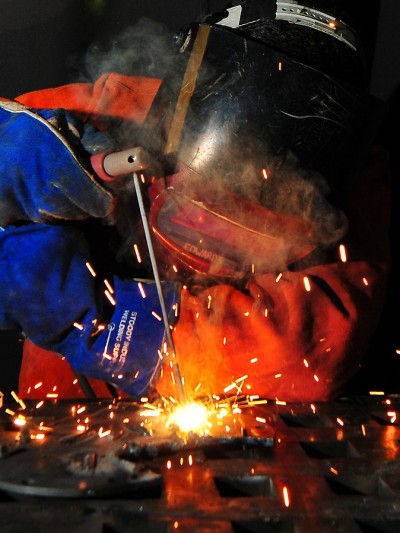 Ein Schweißer in orangener Schutzkleidung, blauen Handschuhen und Helm, der etwas schweißt. Der Hintergrund ist schwarz und Funken sprühen.