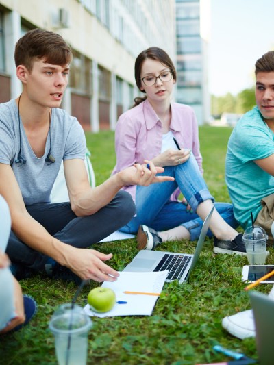 Eine Gruppe von fünf jungen Leuten sitzt bei sommerlichen Temperaturen auf einem Grünstreifen vor einem Gebäude. Die Personen haben ihre Notebooks, Schreibunterlagen sowie Getränke und Snacks dabei. 