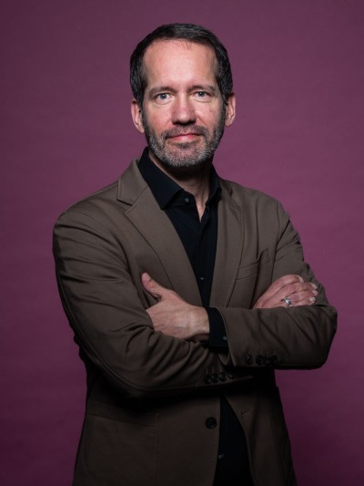 Portrait von Dr. Benny Pock im brauen Anzug vor weinrotem Hintergrund.