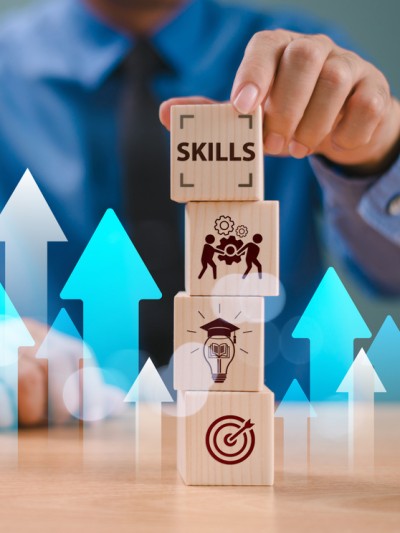 Tisch mit einem Stapel Holzwürfel, auf denen Symbole aus dem Bereich Lernen abgebildet sind. Im Hintergrund ein Anzugträger, der mit seiner Hand die Würfel stapelt. Viele blaue und weiße Pfeile zeigen im Bild nach oben