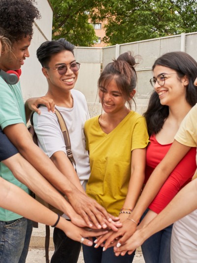 Eine Gruppe aus 8 Jugendlichen in bunten T-Shirts steht im Halbkreis und stapelt ihre Arme aufeinander.