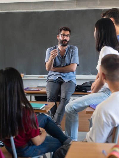 Ein Berufsschullehrer lehnt am Pult und diskutiert mit einigen Schülerinnen und Schülern.
