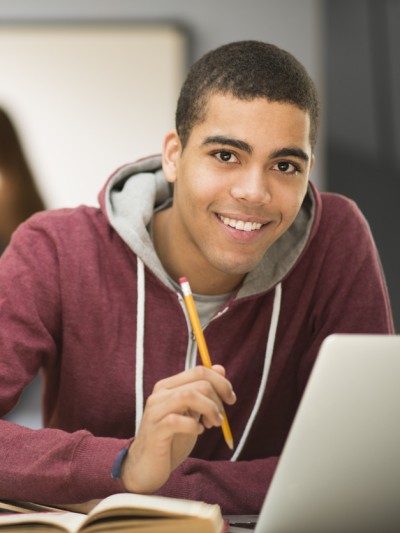 Ein Schüler mit sitzt lächelnd vor seinem Laptop, in der Hand ein Bleistift, auf dem Tisch mehrere aufgeschlagene Bücher.