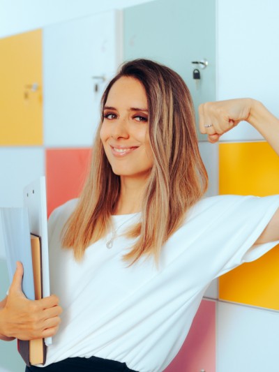 Glückliche Lehrerin vor Schließfächern, die ihren starken Bizeps zeigt und dabei Dokumente in der Hand trägt.