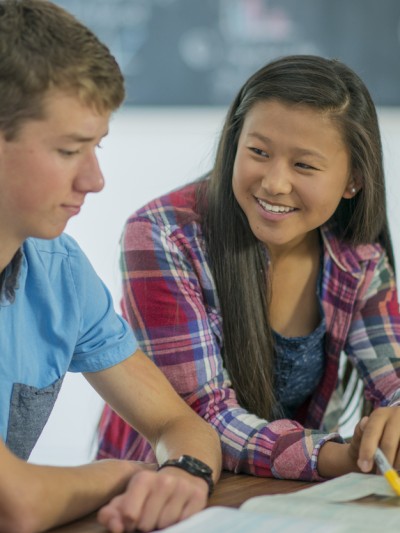 Vier Schülerinnen und Schüler sitzen am Tisch und lernen zusammen. Im Fokus ist eine Schülerin asiatischer Abstammung, die mit dem Stift auf ein Buch zeigt und lächelt.