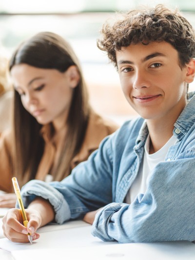Ein Schüler und zwei Schülerinnen sitzen am Tisch und schreiben. Der Junge blickt in die Kamera, die Mädchen blicken auf ihre Papiere vor sich.