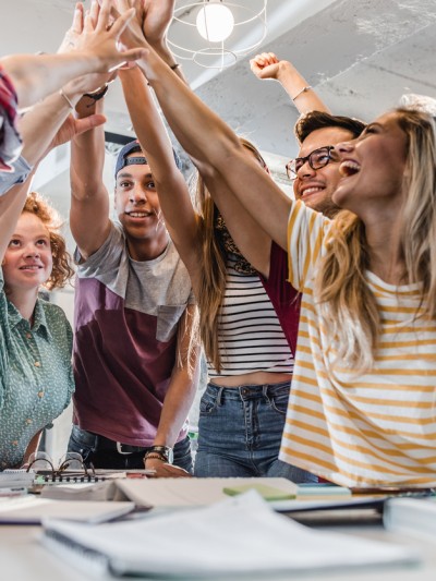 Sechs Jugendliche unterschiedlicher Herkunft und Geschlecht stehen an einem Tisch auf dem Hefte liegen und machen einen Huddle.