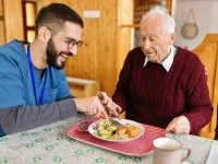 Ein junger Altenpfleger schneidet einem Senior das Essen auf dem Teller klein. Beide wirken entspannt und zufrieden.
