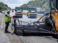 Ein Asphaltbauer, gefolgt von zwei Straßenwalzen, verbaut Flüsterasphalt bei der Sanierung der Autobahn A3 in Nordrhein-Westfalen.